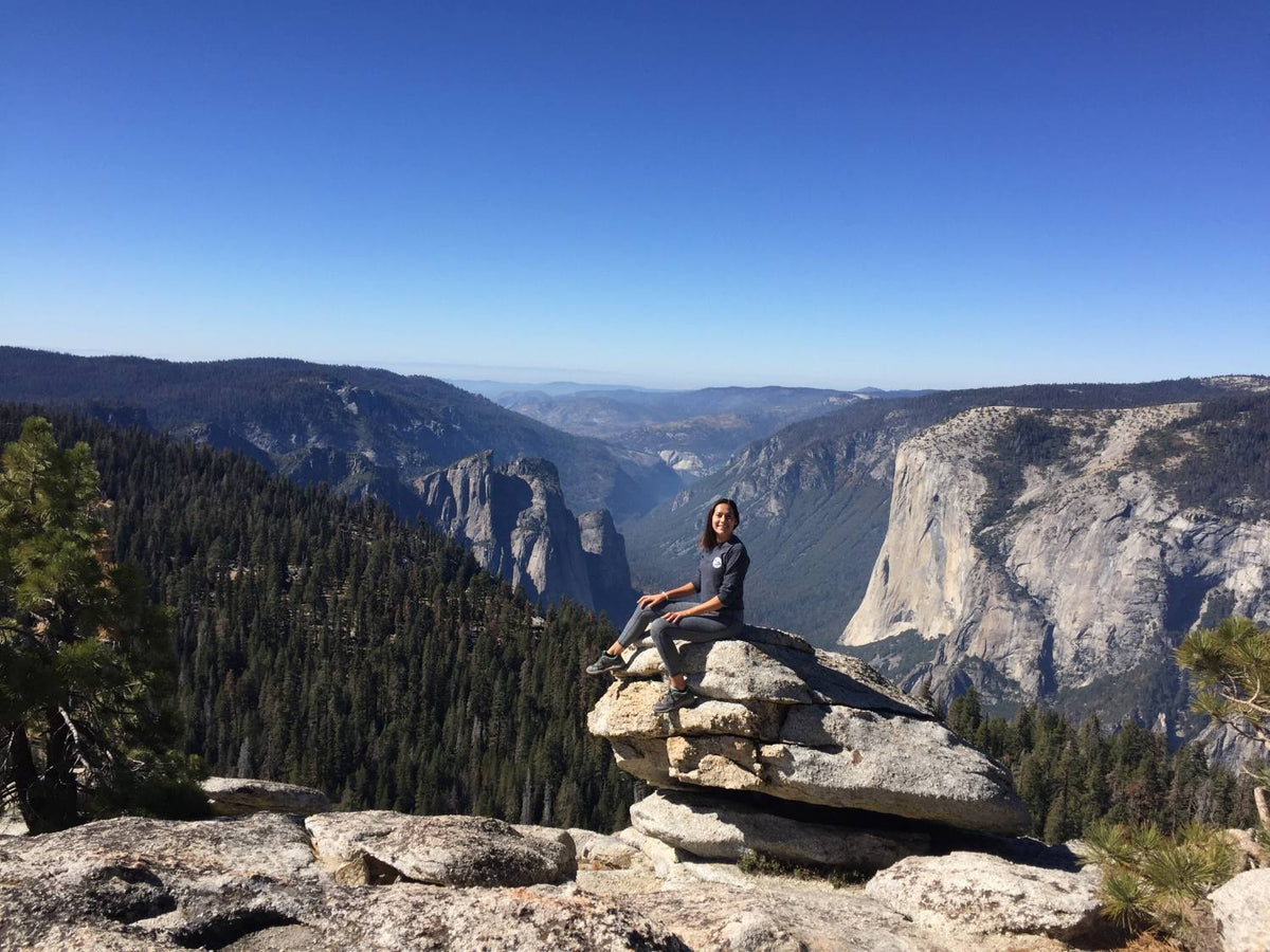 A woman sitting on some rocks and smiling at the camera with mountain ranges behind her
