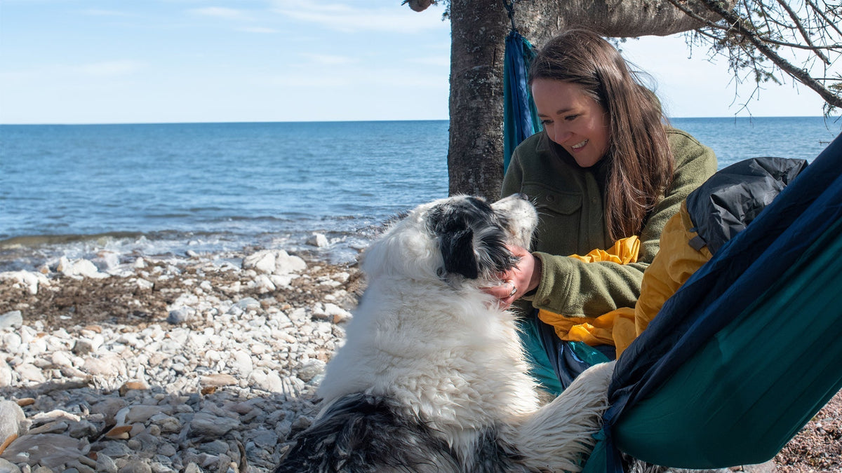 A woman pets and smiles down at her dog while she hammocks on the shore of a beach
