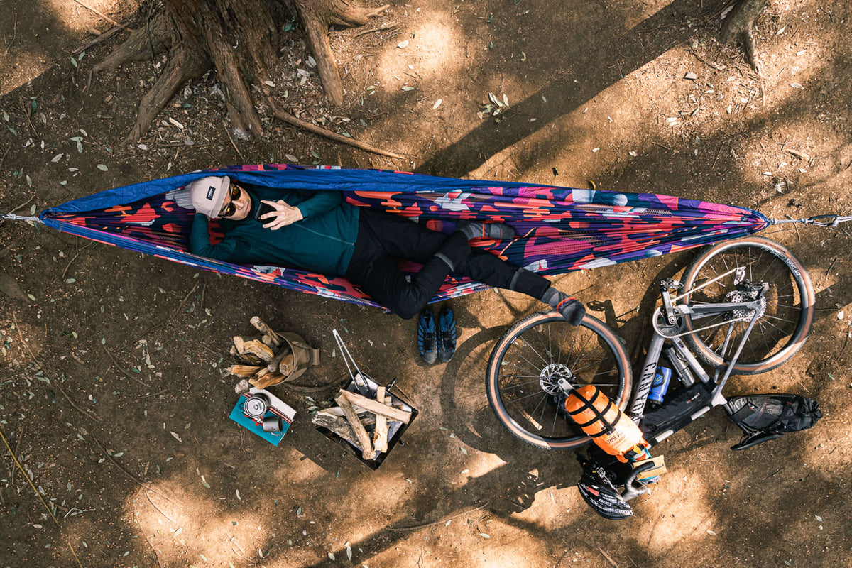 A person laying down in an ENO hammock next to a bike and camping gear