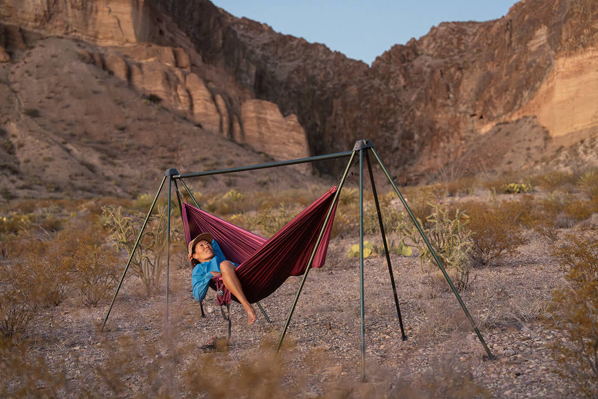 A woman lays in an ENO hammock using the ENO Noamd Hammock Stand. She is in a desert area with mountains behind her