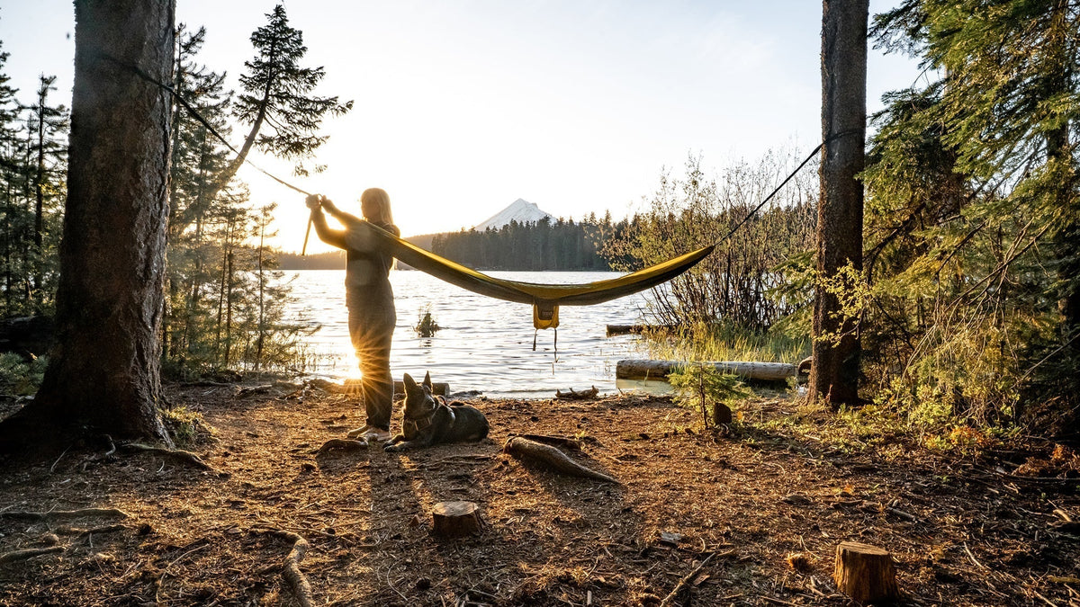 A woman sets up her ENO Hammock between trees in front of a lake and distant mountain view.
