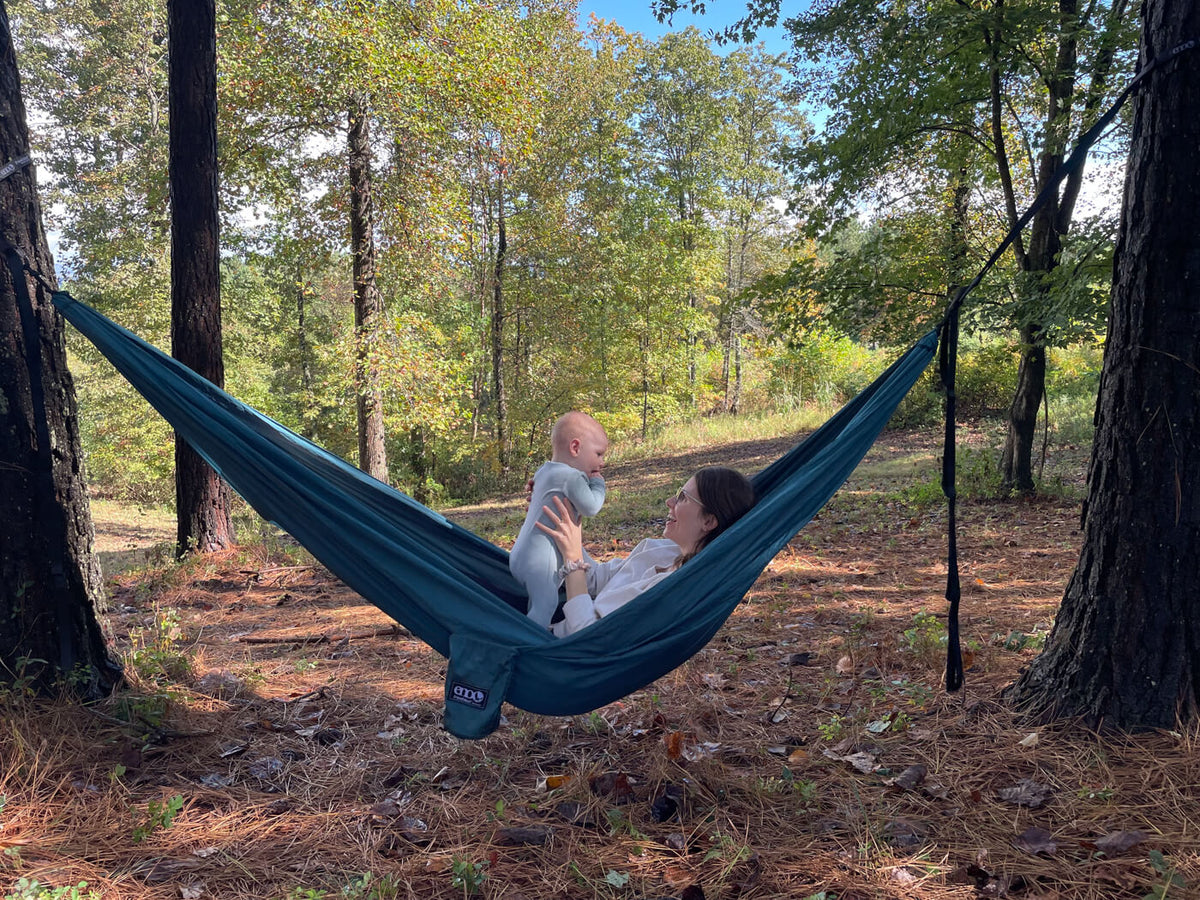 A woman holds a baby while in an ENO TravelNest Hammock. They are in the woods.