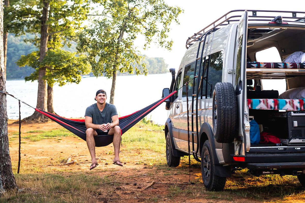 A man sits in an ENO Hammock that is hung from a tree and van with a body of water behind him