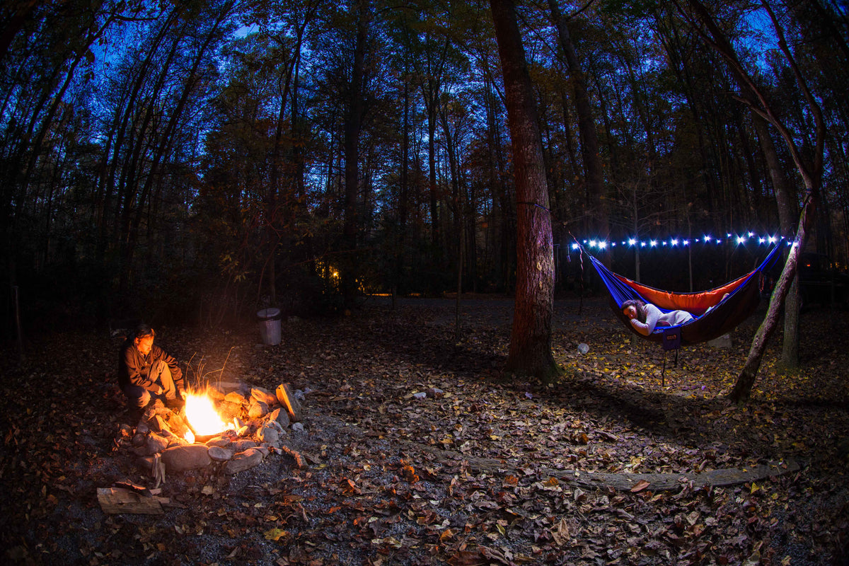 A girl in an ENO hammock with ENO Twilights above head watching a guy attend to a campfire