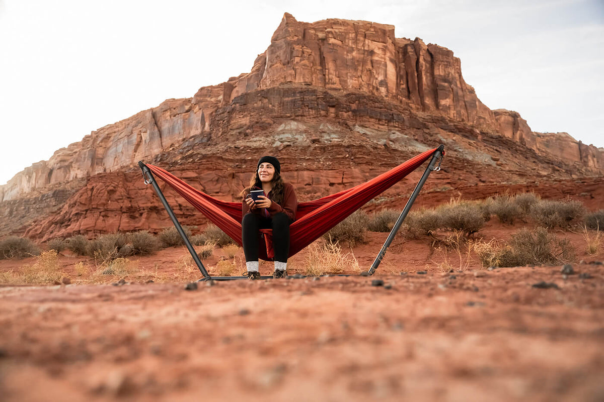 A woman sits in her ENO Canyon/Coral DN hammock using the Parkway Adjustable Hammock Stand while in the middle of a desert of red rocks.