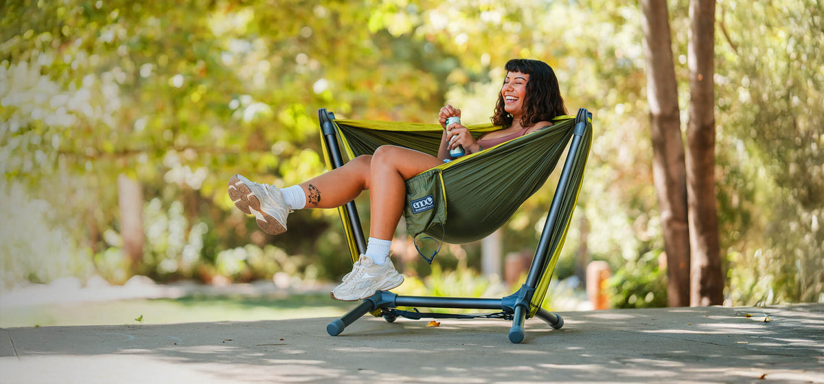 Woman sits back, kicking a leg up with beverage in hand, in the ENO Parklite Hammock Chair Stand with DoubleNest Hammock in front of lush green backdrop.