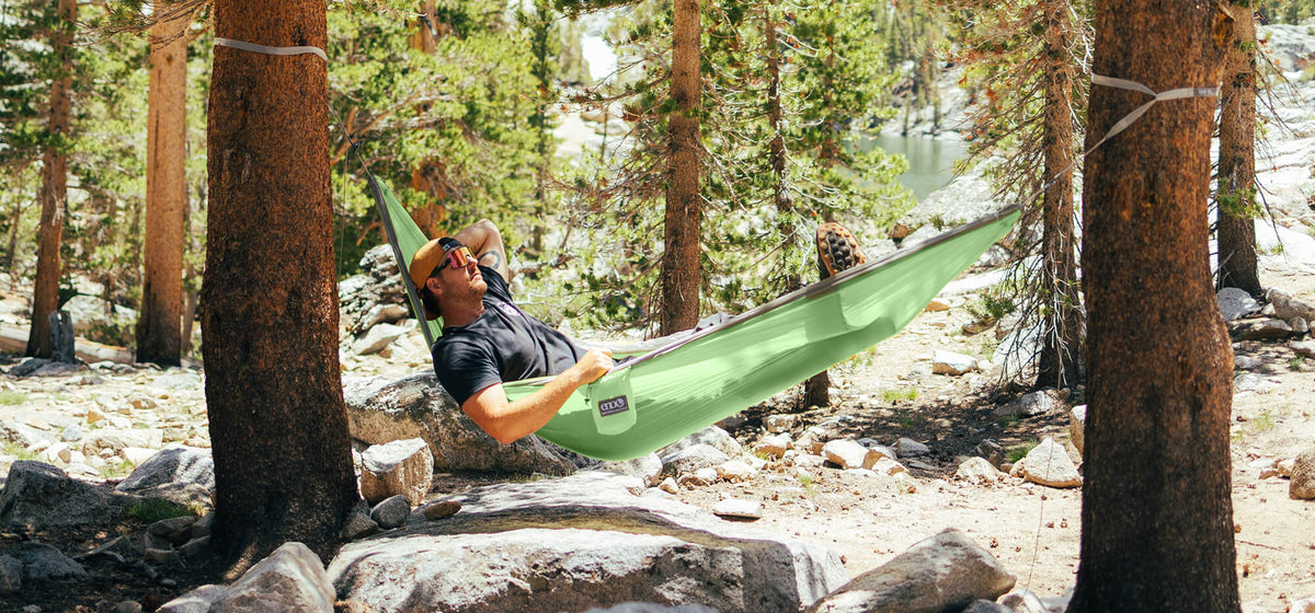 Person relaxing in a SuperSub Ultralight Hammock in Cactus Steel colorway between two trees in a forest setting.