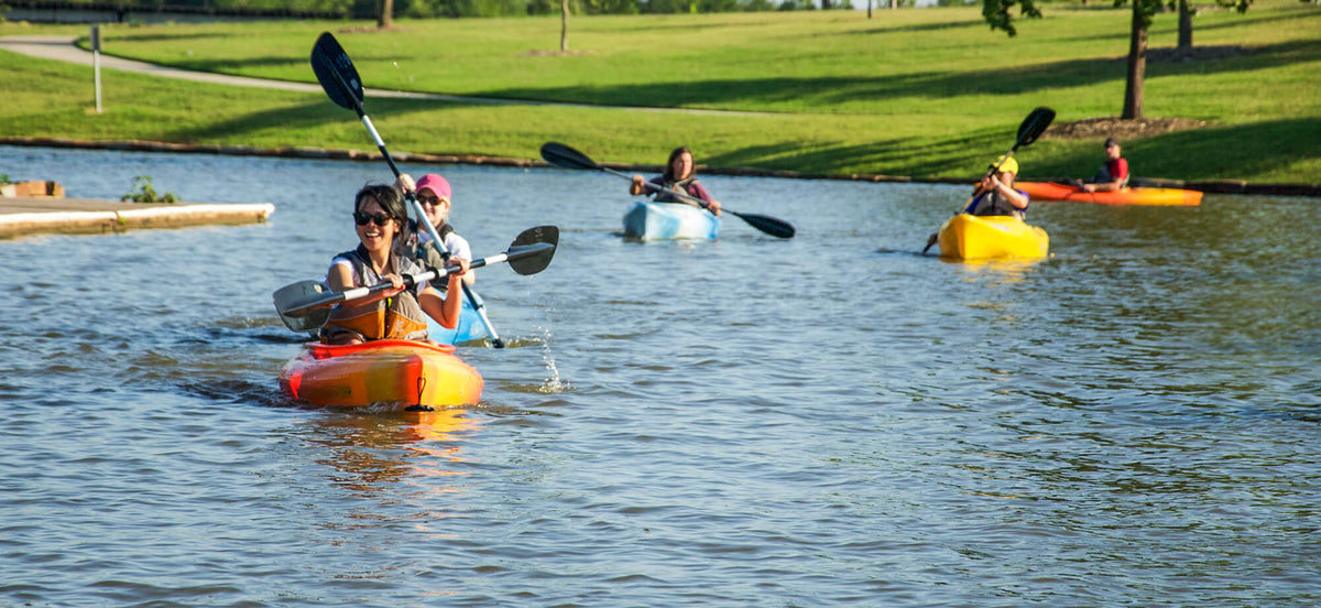 People kayaking on a calm body of water with green grass and trees in the background