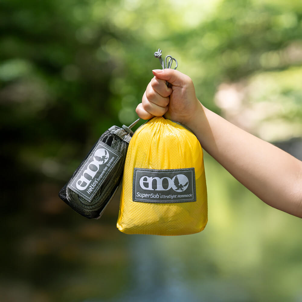 A person holds a packed ENO SuperSub Ultralight Hammock and Helios Hammock Straps in the air with a soft green forest in background.