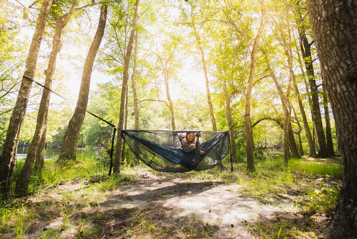 A girl in an ENO Hammock using an ENO Guardian DX Bug Net. She is in the woods with the sun shining through the trees.