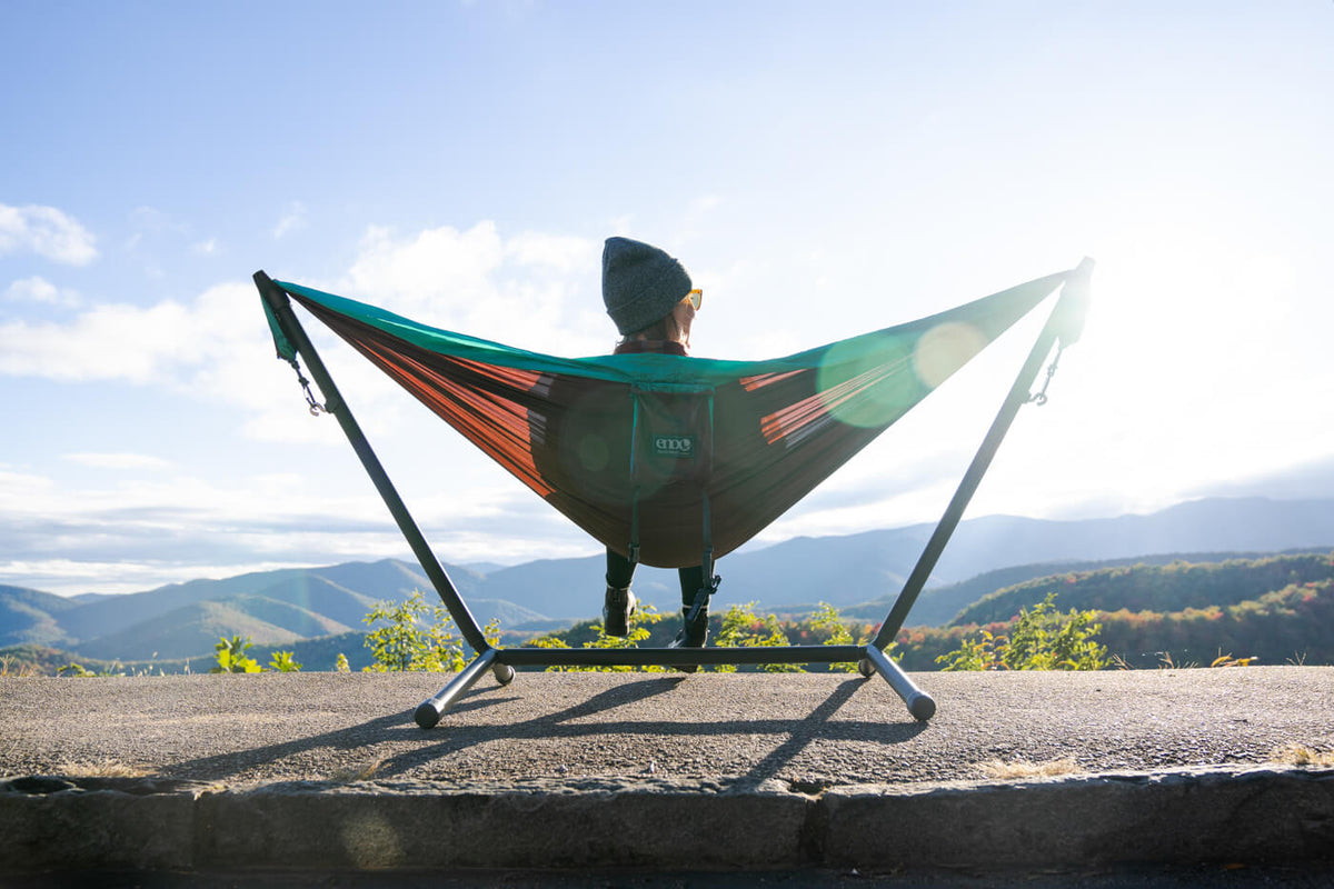A woman sitting in an ENO hammock in the ENO Parkway Adjustable Hammock Stand while at a Blue Ridge Parkway lookout of the mountains