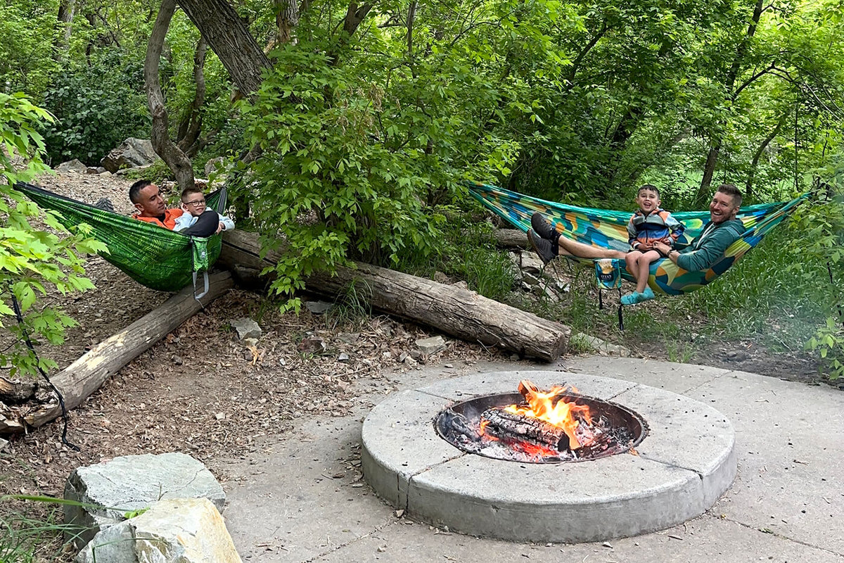 Four people in 2 ENO DoubleNest Print Hammocks in the woods with a firepit in front of them