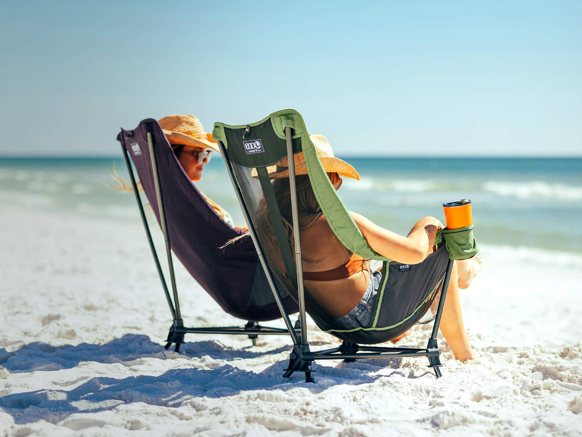 Two women lounging in low camp chairs on the beach one in mesh and one solid . Cope on screen reads: Redefining relaxation with our new Lounger DL Chairs