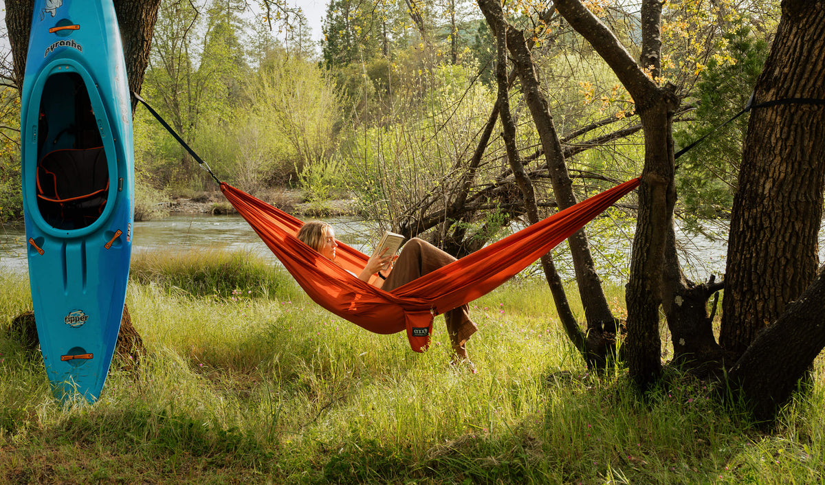 A young woman lays back in an ENO TravelNest Hammock + Straps Combo in new Canyon color while reading a book in a lush green landscape with river in background. 