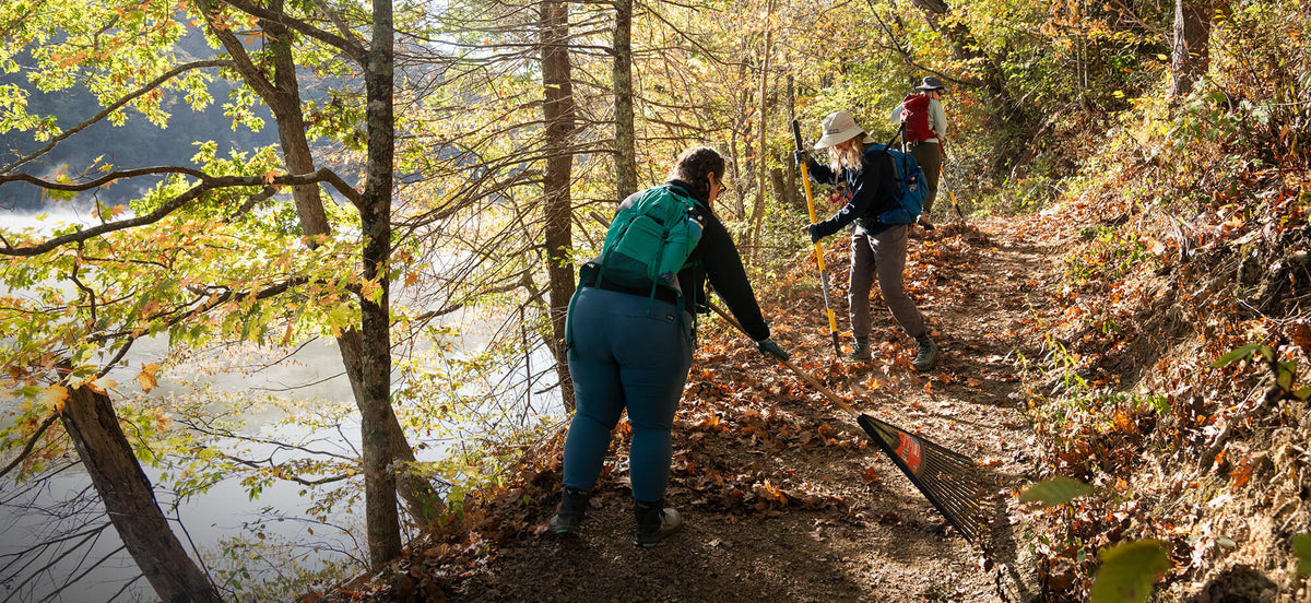 Three hikers on a trail by a lake with trees and autumn foliage.