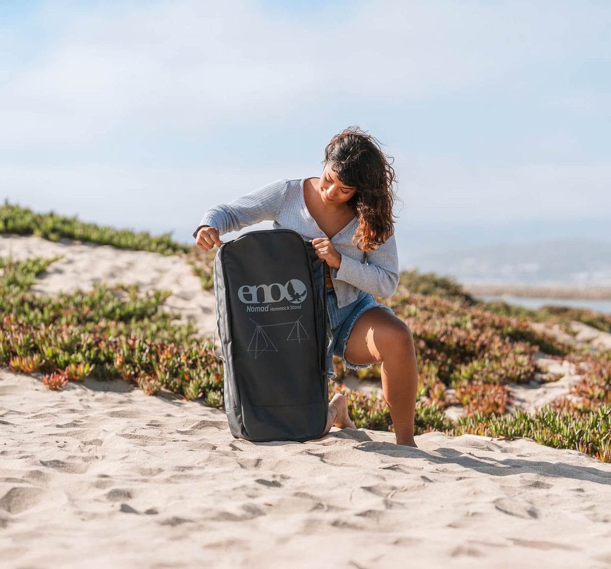 Person sitting on a sandy beach opening a packed ENO Nomad Hammock Stand custom backpack.
