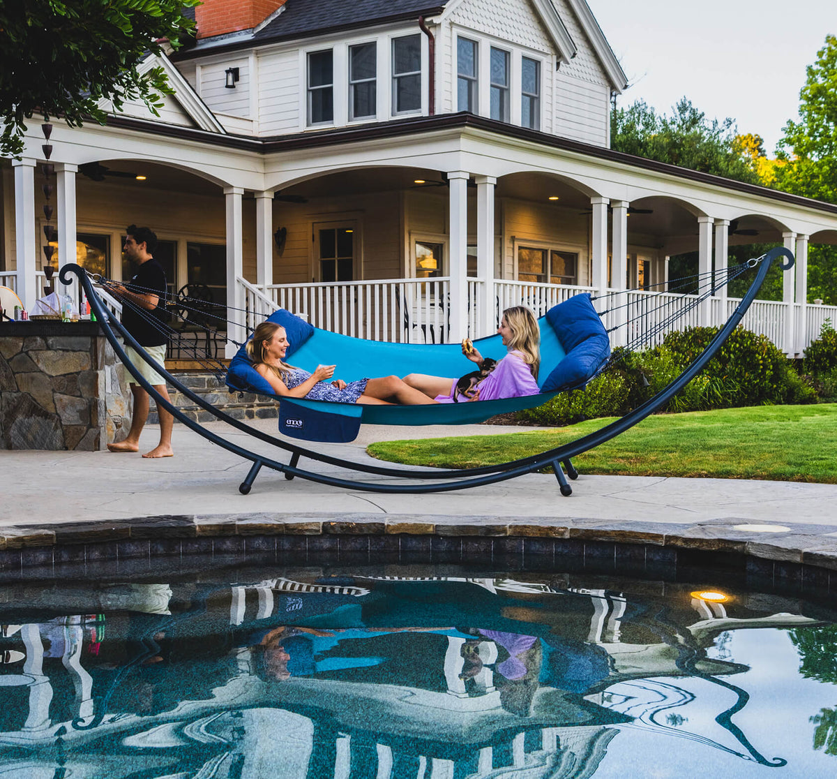 Two people laying in a ENO SuperNest SL Hammock set up on an ENO SuperNest XL Hammock Stand by a pool with a house in the background.