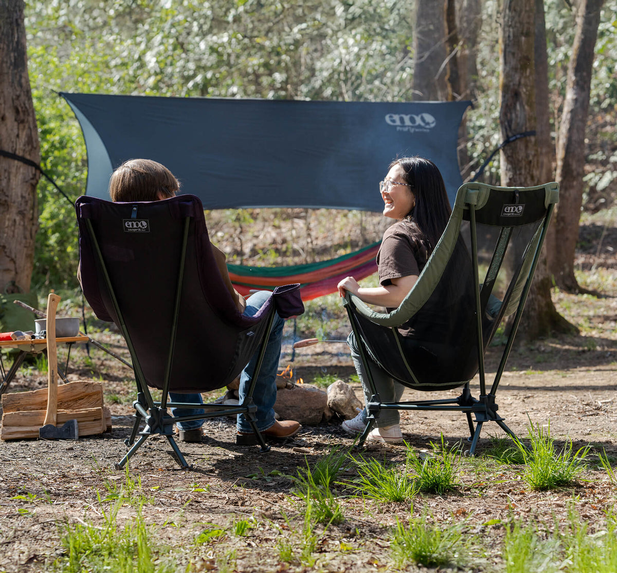 Two people sitting in ENO Lounger DL Chairs with a hammock and tarp set up in the background of a forest setting.