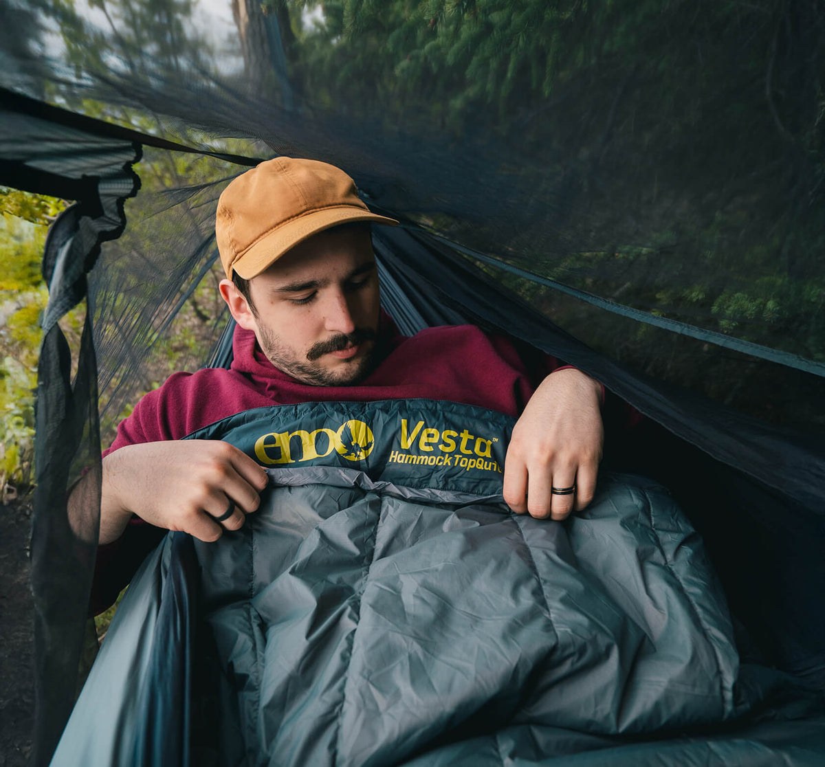 Man sitting in a hammock with a Vesta Hammock TopQuilt, surrounded by greenery and enclosed within an ENO Bug Net.