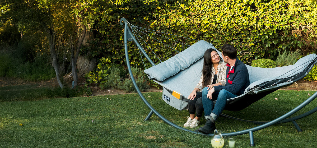 Two people sitting on a SuperNest Hammock set up on a SoloPod XL Hammock Stand in a garden setting.