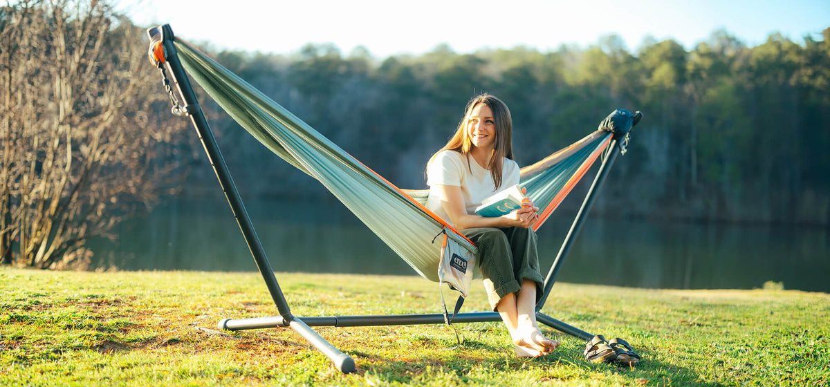 A person sitting in a ENO DoubleNest Hammock on a Parkway Adjustable Hammock Stand with a book in a like setting.
