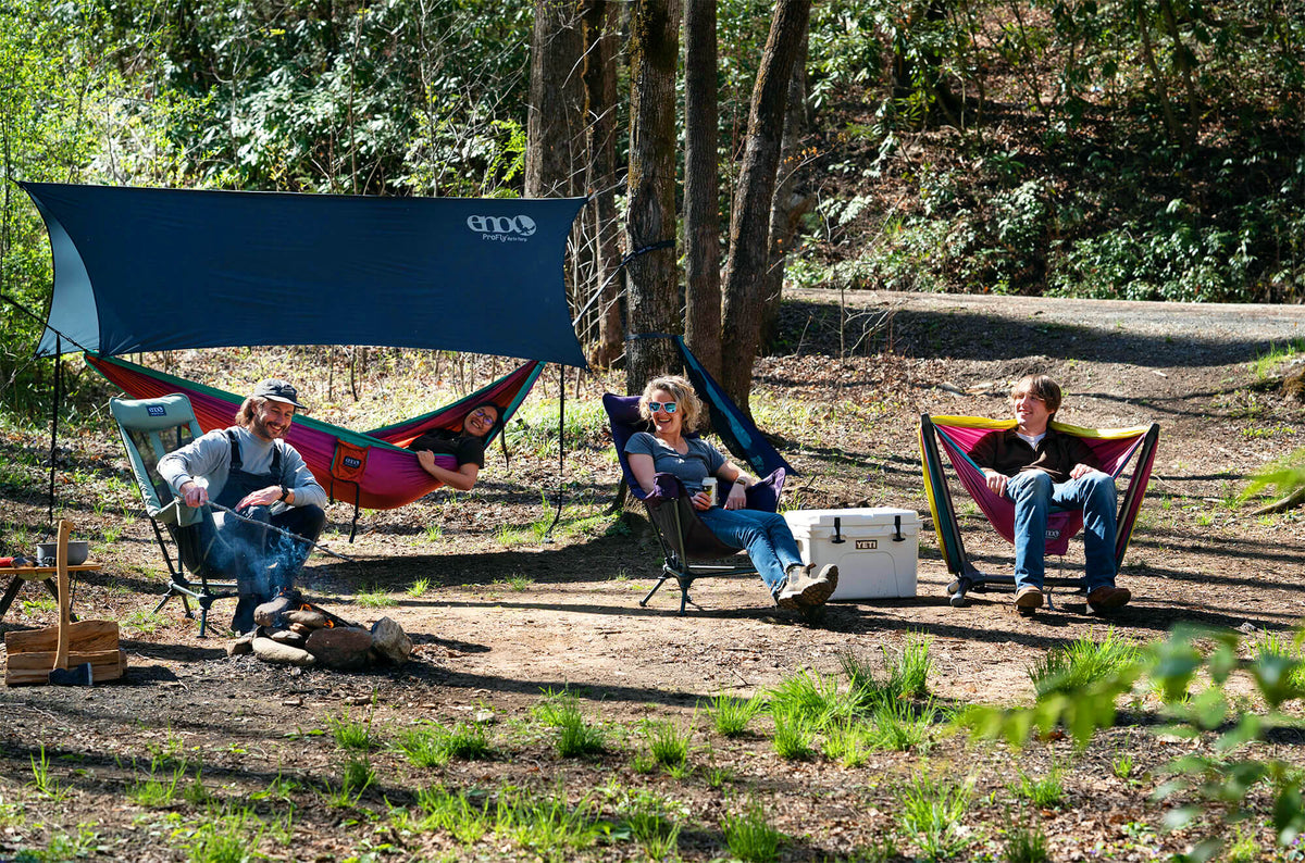 A group of campers sit fireside laughing in a range of ENO products including a Lounger DL Chair, Lounger DL Mesh Chair, ProFly Rain Tarp, DoubleNest Hammock Print, and DoubleNest Hammock setup in a Parklite Hammock Chair Stand.