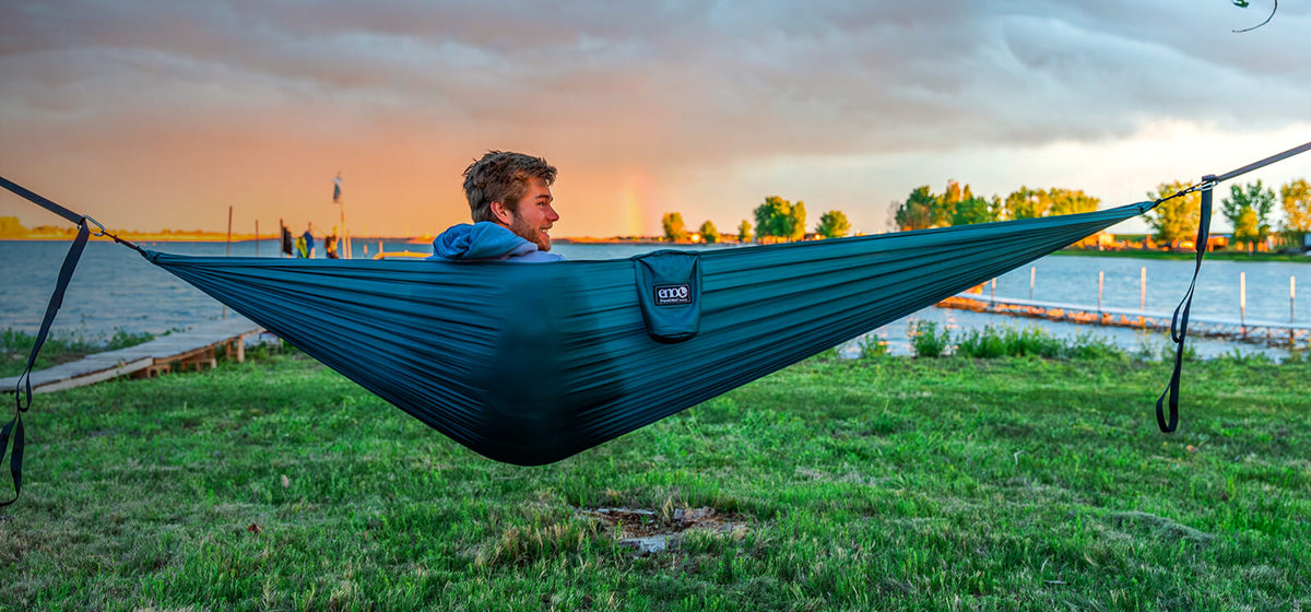 A person sits in a ENO TravelNest Hammock + Straps Combo while looking back in front of a lake with rainbow on the skyline. 