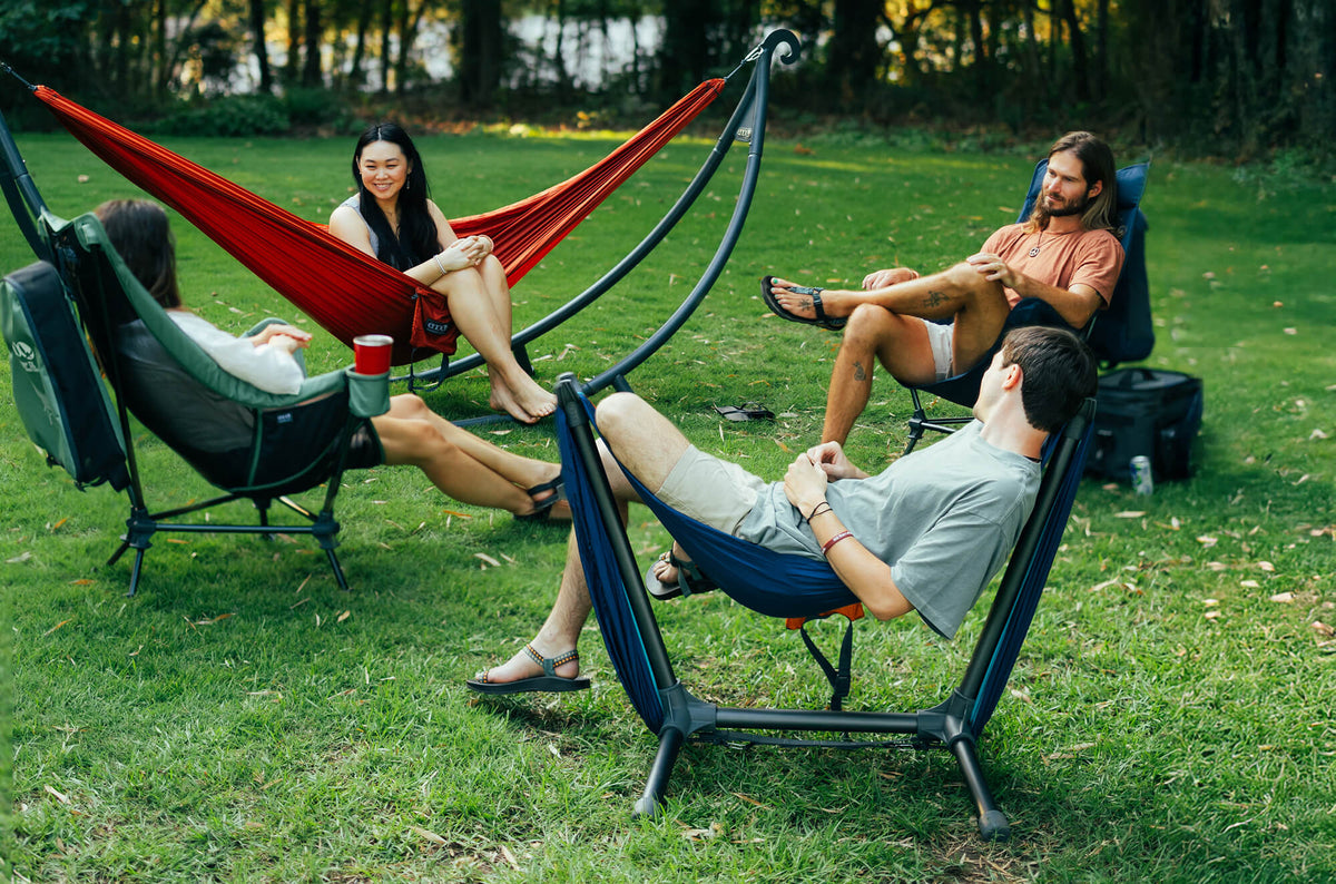 A group of friends sit in a circle of ENO products consisting of ENO Lounger DL Mesh and Solid chairs, a TravelNest Hammock set up on a SoloPod Hammock Stand and a DoubleNest Hammock set up on a Parklite Hammock Chair Stand in a backyard environment.