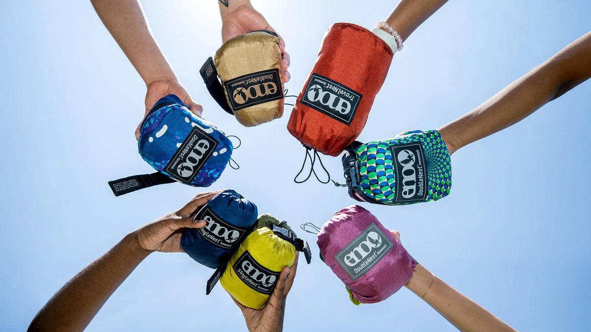 Multiple hands holding a variety of colorful packed ENO Hammock Stuff Sacks against a clear blue sky