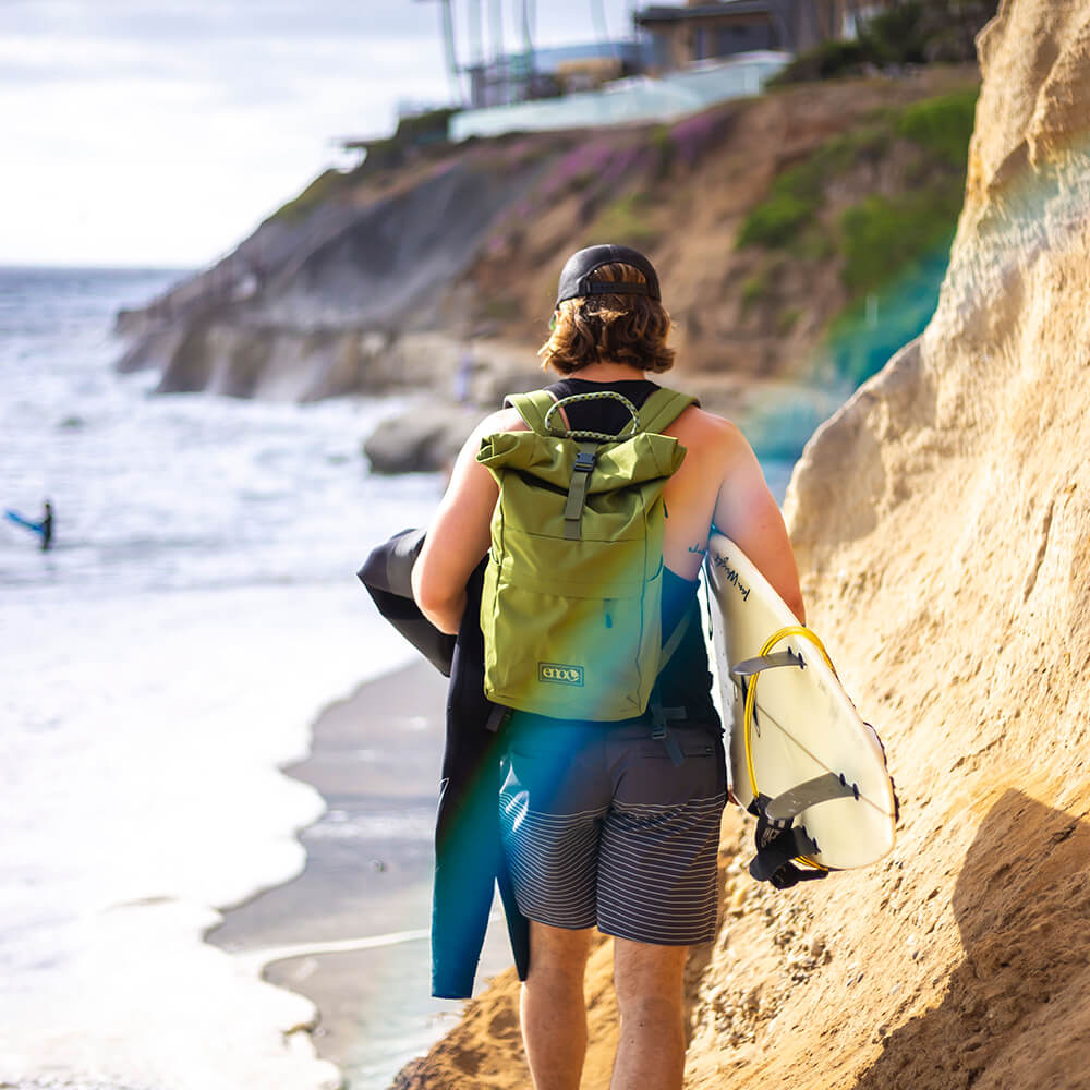 A person walk along a Cliffside trail towards the ocean while carrying a surf board with an ENO Roan Rolltop Pack on their back.  