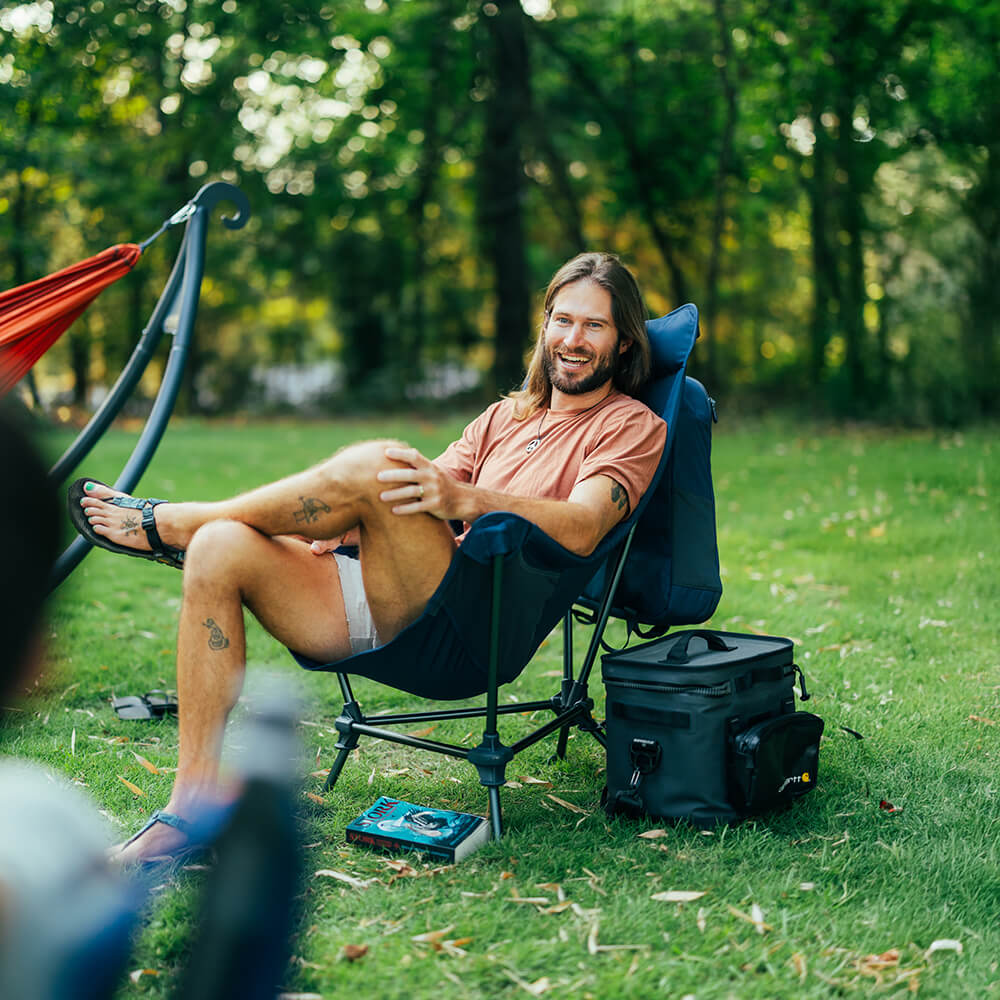 A young man sits, talking with a group of friends, in an ENO Lounger DL Chair within a lush green back yard environment. 