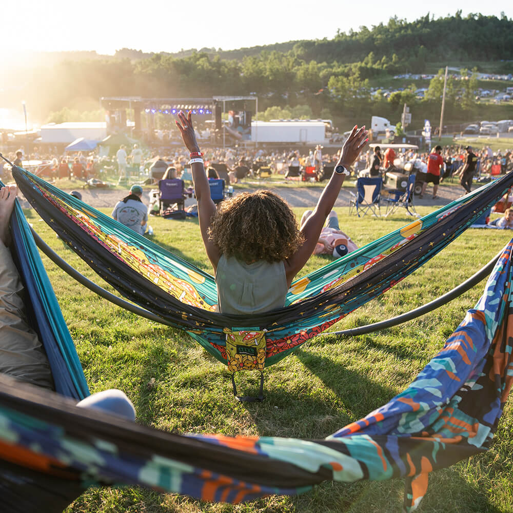 A woman cheers on a band with their hands in the air while sitting in an ENO DoubleNest Hammock Print at a music festival.