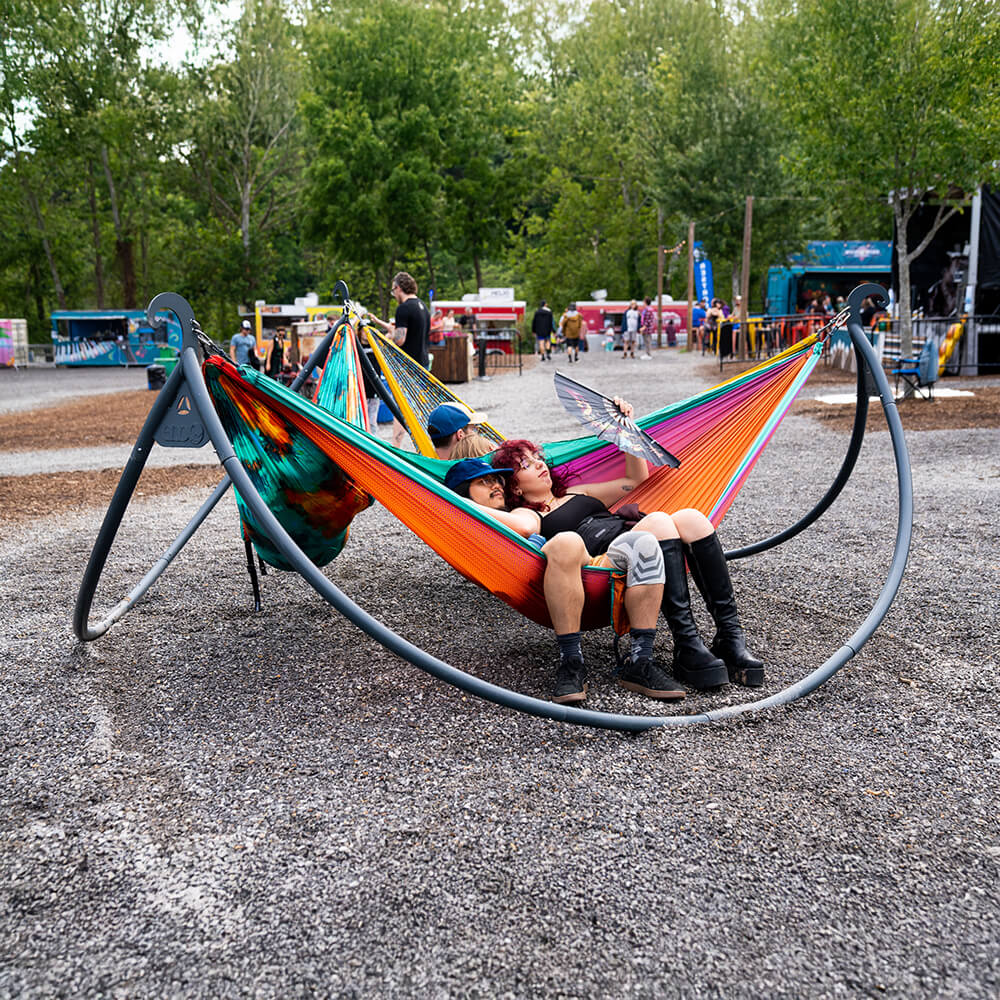 A group of friends sit back in three separate DoubleNest Hammock Prints set up on the ENOpod Triple Hammock Stand at a music venue. 