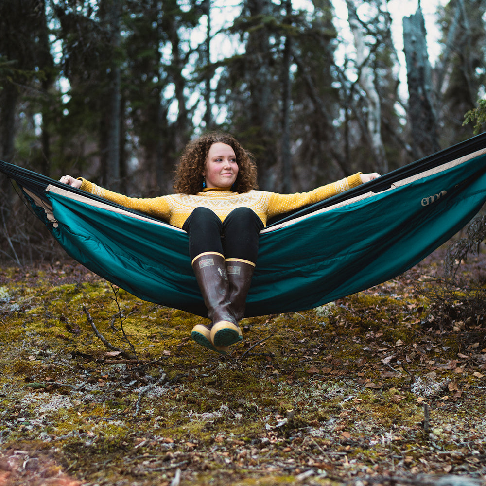 A woman sits up in their ENO DoubleNest Hammock with Ember UnderQuilt underneath in an old growth forest with moss underneath.
